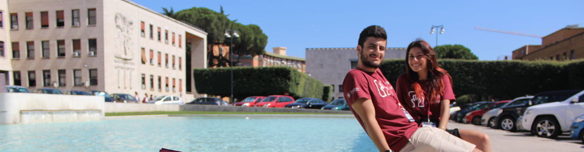 Uno studente e una studentessa seduti sulla fontana della minerva con il cartello #Io scelgo Sapienza